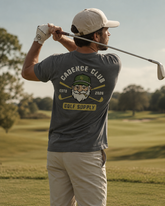 A man on a golf course wearing a dark gray t-shirt with a 'Cadence Club Golf Supply' graphic on the back, paired with khaki pants and a beige cap.