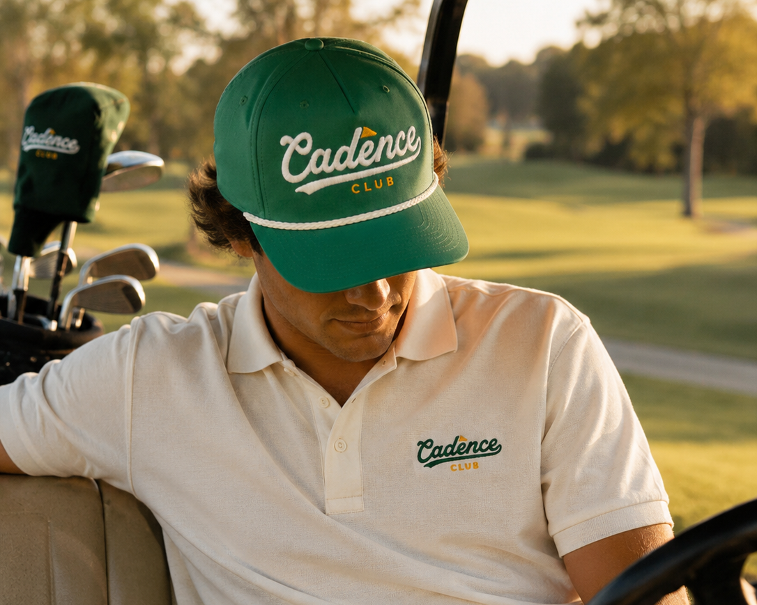 Man sitting in a golf cart wearing a green 'Cadence Club' cap and white polo shirt on a golf course.