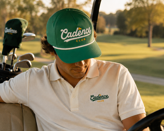 Man sitting in a golf cart wearing a green 'Cadence Club' cap and white polo shirt on a golf course.