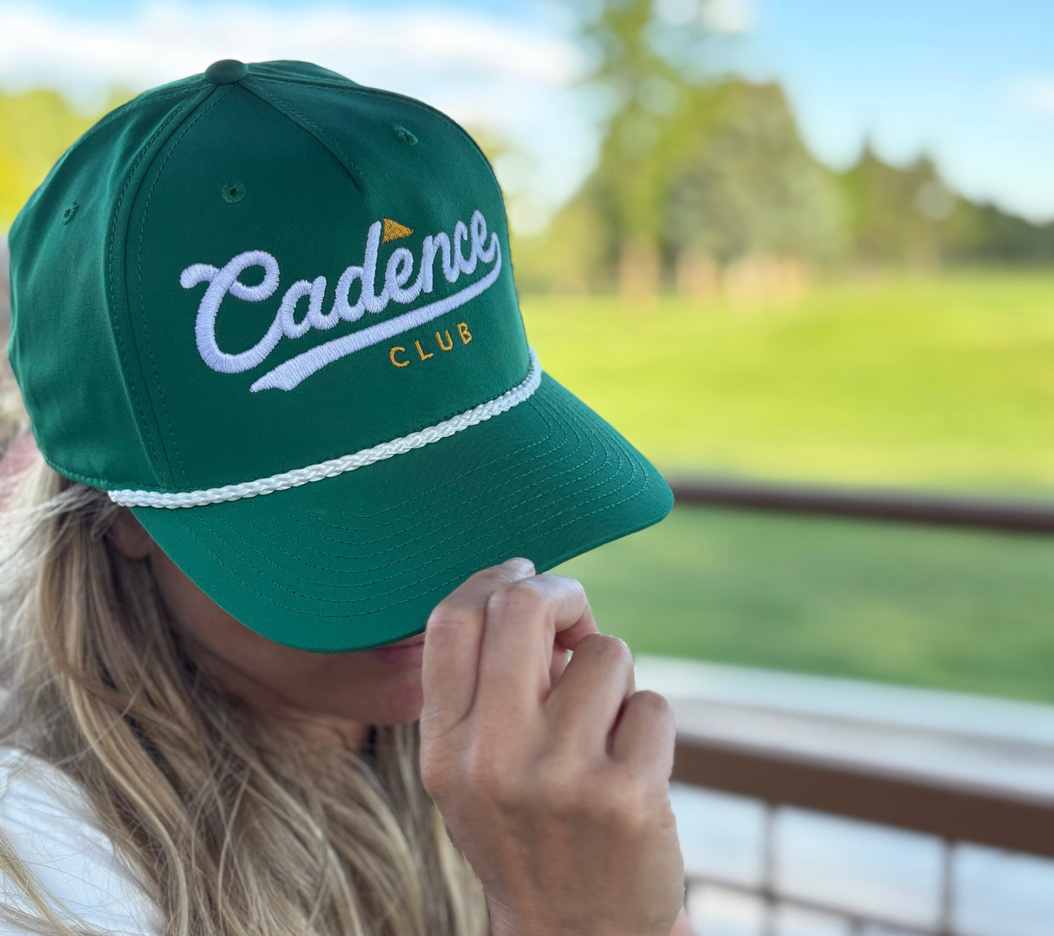 A woman wearing a green baseball cap with 'Cadence CLUB' embroidered in white and yellow, photographed outdoors on a patio with a blurred golf course background.