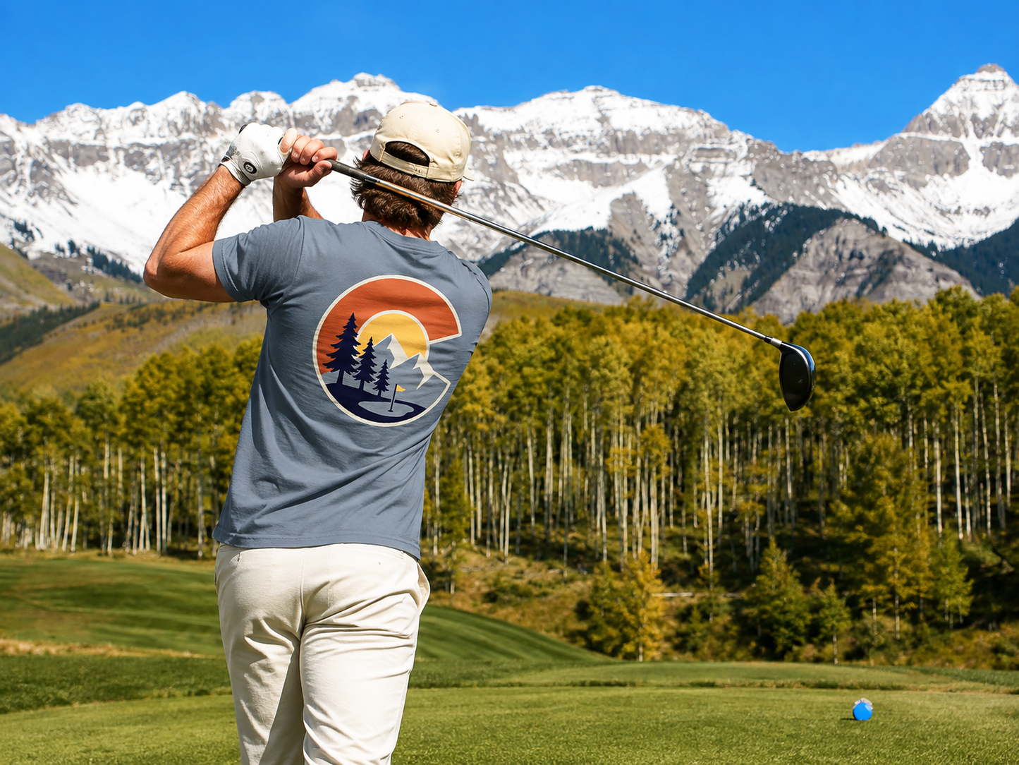 A man on a golf course wearing a gray t-shirt with a large circular sunset and pine tree graphic on the back, paired with off-white pants and a beige cap, with snow-capped mountains and green trees in the background.