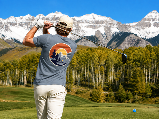 A man on a golf course wearing a gray t-shirt with a large circular sunset and pine tree graphic on the back, paired with off-white pants and a beige cap, with snow-capped mountains and green trees in the background.