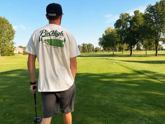 A man on a golf course seen from behind, wearing a white t-shirt with a green 'PinHigh' logo and a black cap, holding a golf club.