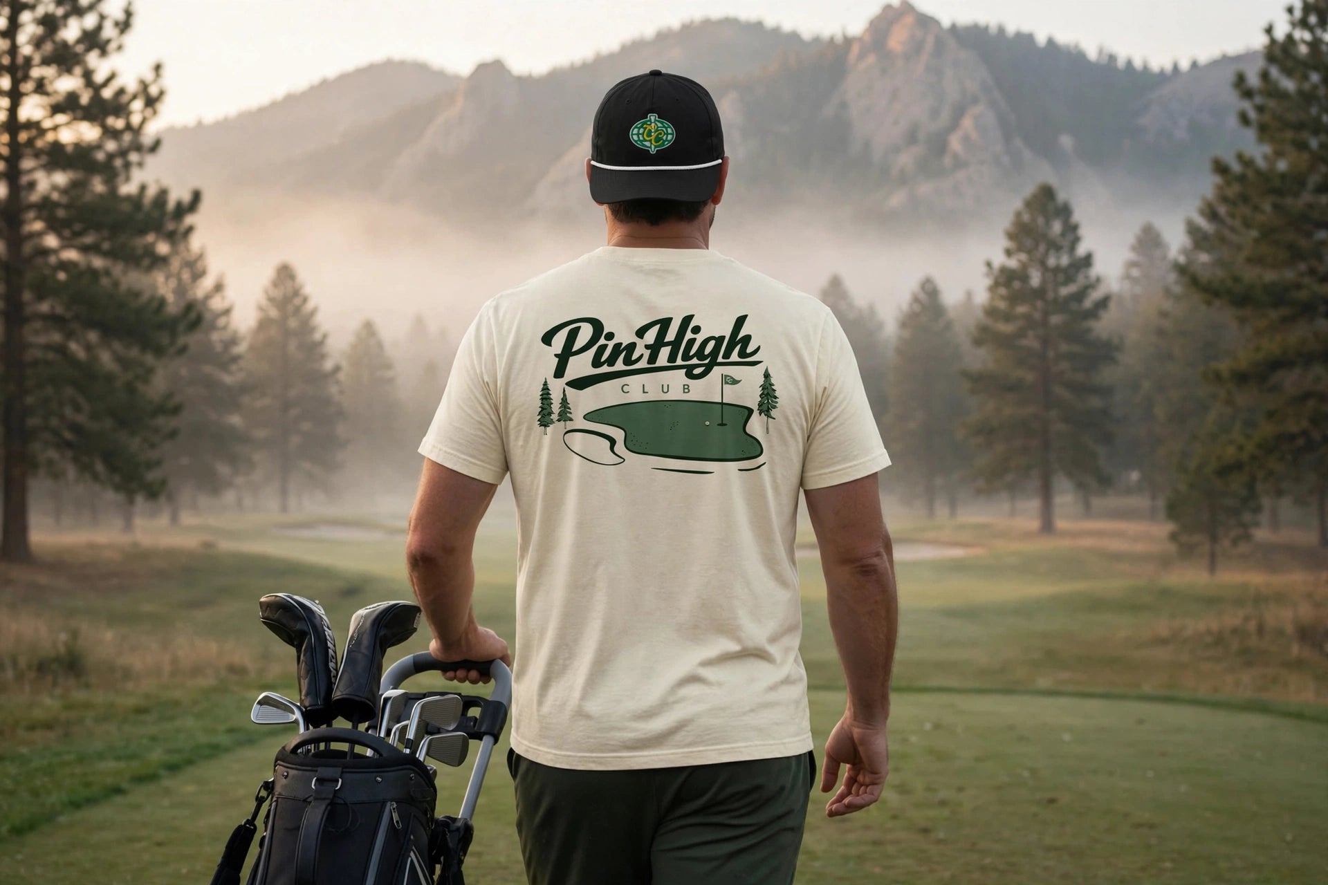 A man seen from behind on a misty golf course, wearing a cream-colored t-shirt with a green PineHigh Club logo on the back, a black baseball cap, and carrying a golf bag.