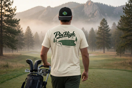 A man seen from behind on a misty golf course, wearing a cream-colored t-shirt with a green PineHigh Club logo on the back, a black baseball cap, and carrying a golf bag.