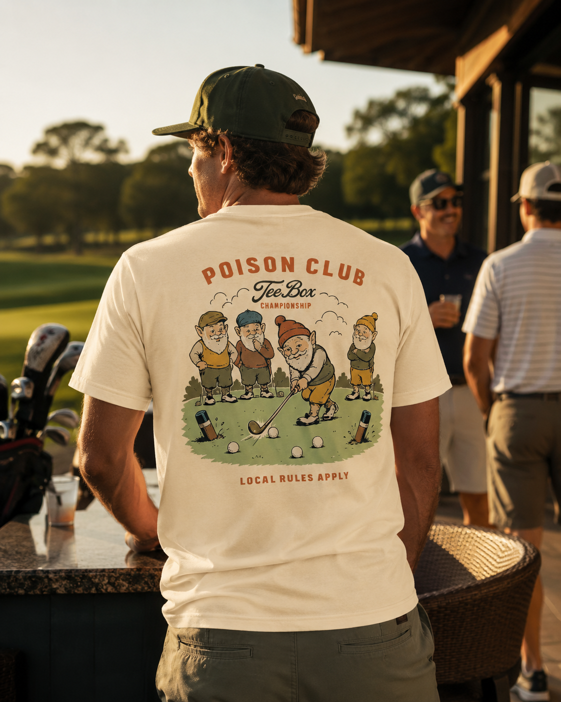 A man standing on a golf course patio wearing a cream-colored t-shirt with a large back graphic that reads 'POISON CLUB Tee Box GOLF CLUB' and shows cartoon characters playing golf. Other people and a golf cart are visible in the background.