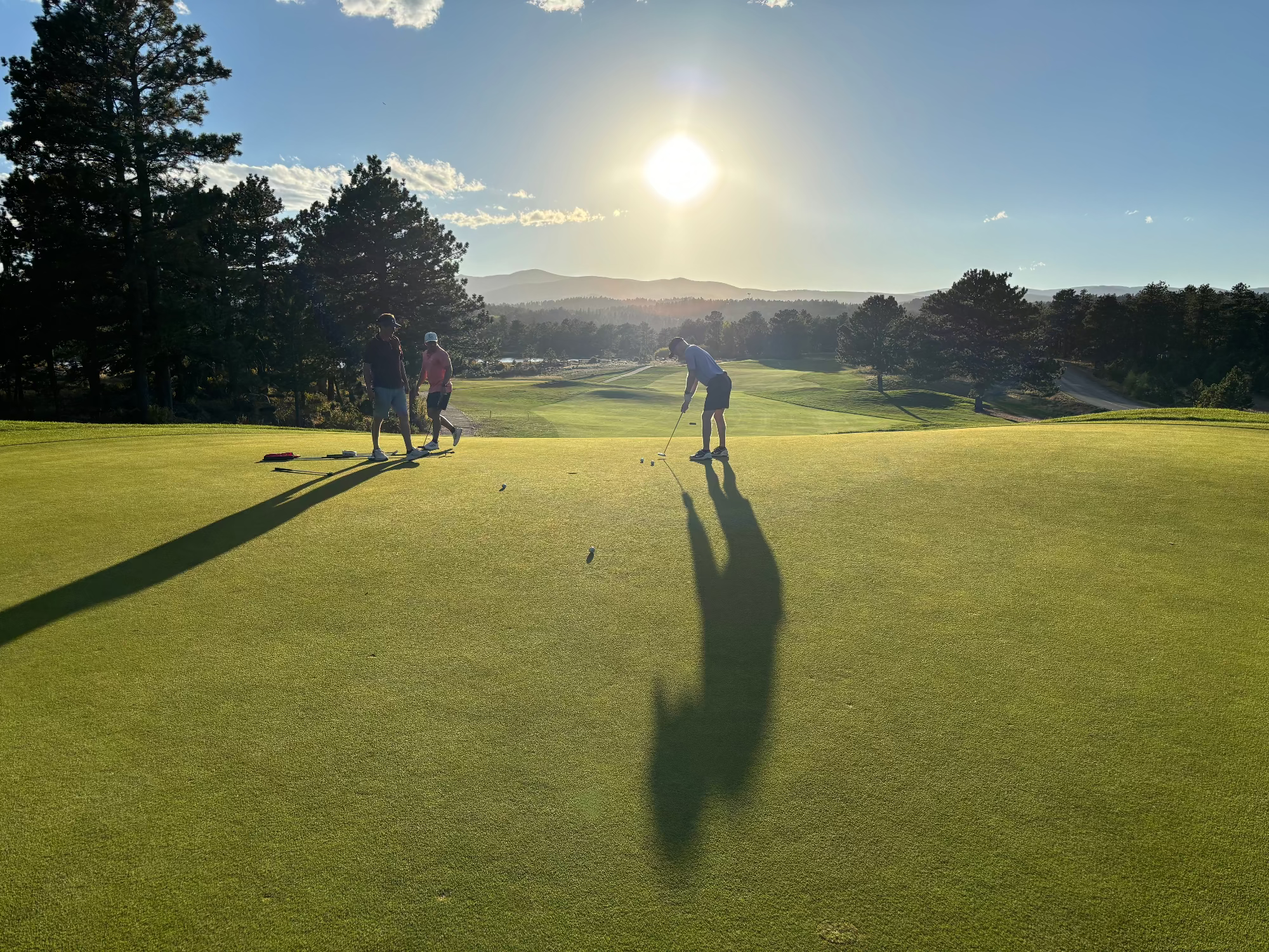 A scenic outdoor golf course at sunset with several people on the green, long dramatic shadows stretching across the grass, trees, and mountains in the background under a bright sun.