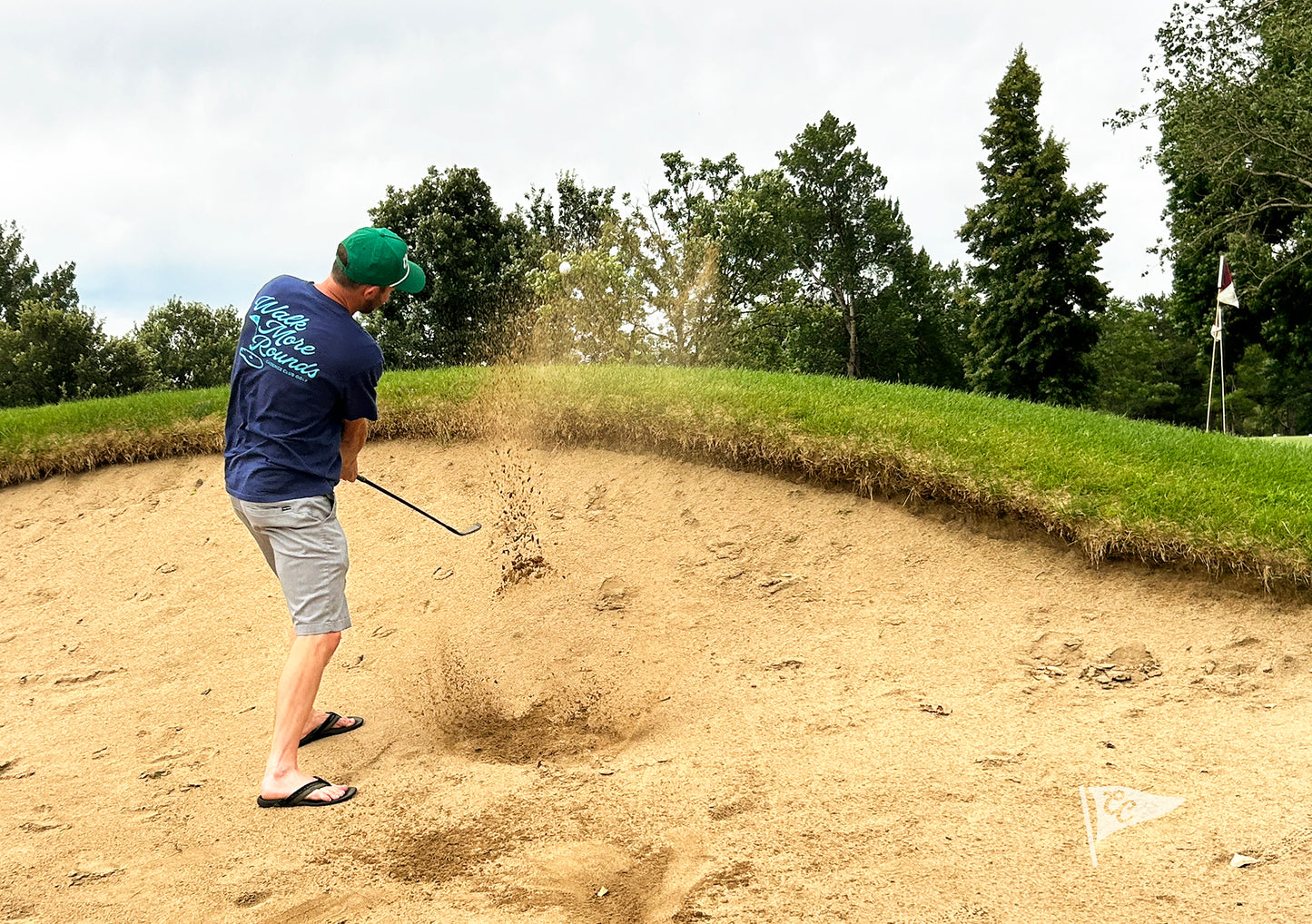 A man in a golf bunker wearing a navy blue shirt, gray shorts, and a green cap, mid-swing with sand flying up. Trees and a grassy hill are visible in the background.