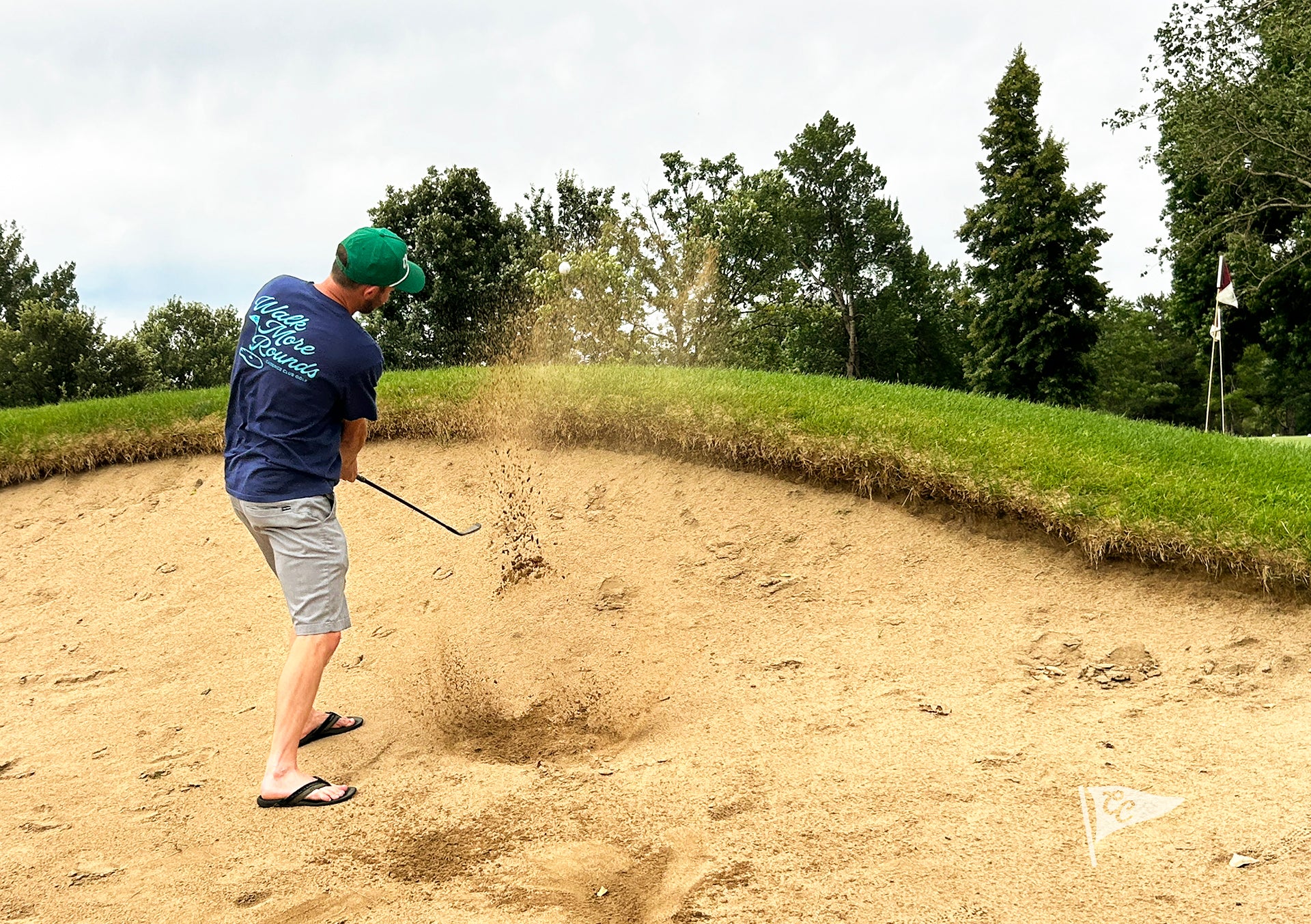 A man in a golf bunker wearing a navy blue shirt, gray shorts, and a green cap, mid-swing with sand flying up. Trees and a grassy hill are visible in the background.
