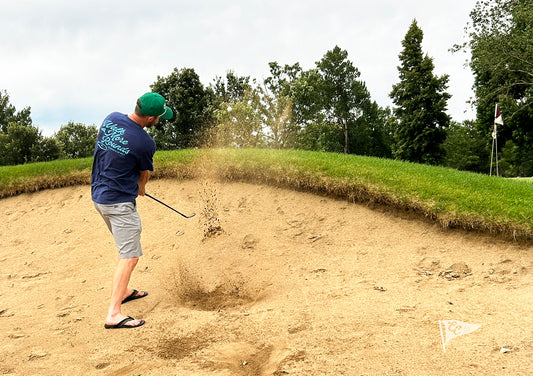 A man in a golf bunker wearing a navy blue shirt, gray shorts, and a green cap, mid-swing with sand flying up. Trees and a grassy hill are visible in the background.