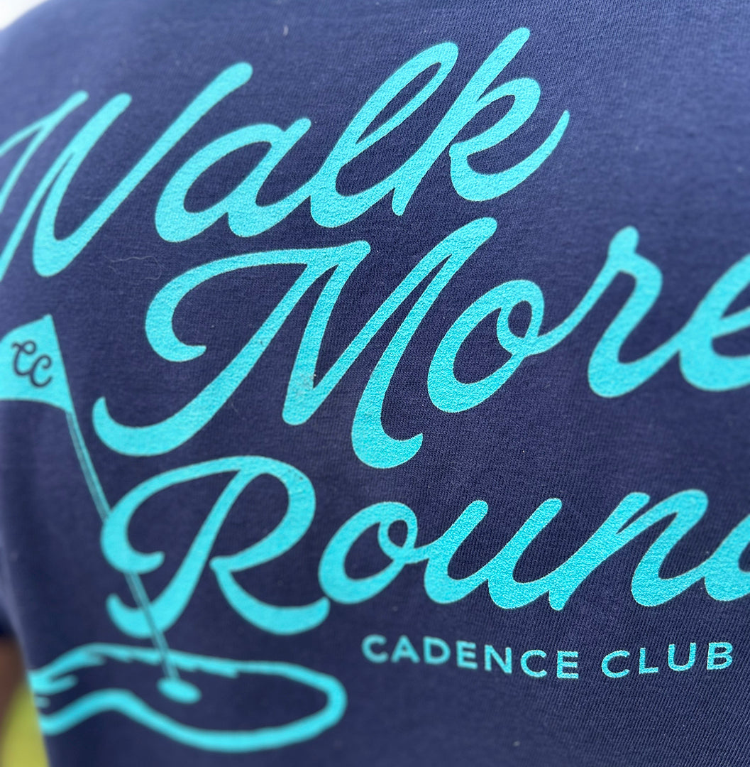 Close-up of a navy blue fabric surface with large light aqua script reading 'Walk More Rounds' and smaller text 'CADENCE CLUB' plus a golf flag graphic.