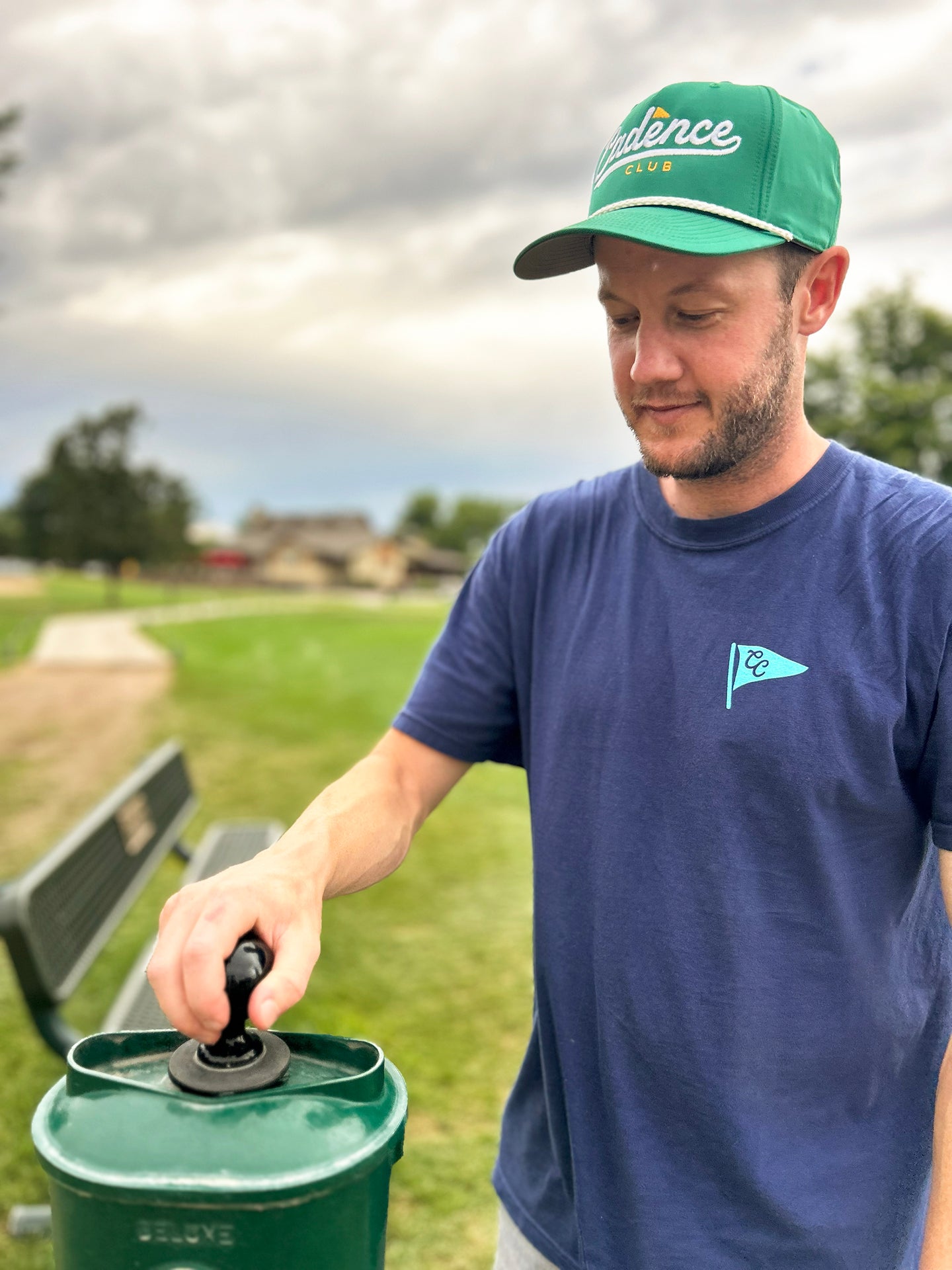 A man wearing a green 'Pace' baseball cap and a navy blue t-shirt with a small light blue flag logo, opening the lid of a green metal container outdoors on a grassy field.