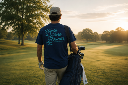 Man walking on a golf course with a bag of clubs, wearing a blue t-shirt with text.